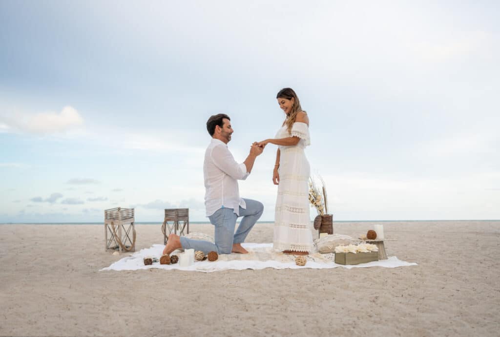 Loving couple getting engaged on a romantic picnic at the beach and him on his knees while he puts the princess cut diamond ring on her finger.