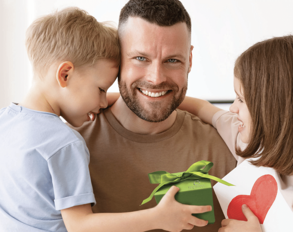 A joyful moment where a smiling man is being celebrated by two children. The man, dressed in a tan T-shirt, holds a young boy in a light blue shirt, while a young girl in a pink shirt, holding a handmade card with a red heart, presents him with a gift from Brinker's Jewelers wrapped in green paper and adorned with a green ribbon. The image conveys warmth, affection, and the joy of giving and receiving, fitting for a special occasion like Father's Day.