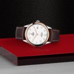 Tudor watch on red surface, featuring a silver case, rose gold bezel, and brown leather strap.