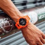 Vibrant orange G-Shock watch on muscular wrist against textured graffiti background.