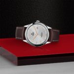 Tudor wristwatch with silver case, textured dial, and brown leather strap on a red base.