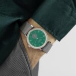 Stylish wristwatch with green dial and gray strap on a persons wrist in a green shirt.