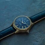 Elegant gold watch with black dial and crocodile leather strap on a textured slate background.