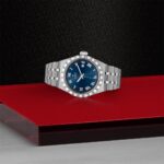 Tudor luxury watch on red surface with blue dial and stainless steel bracelet.