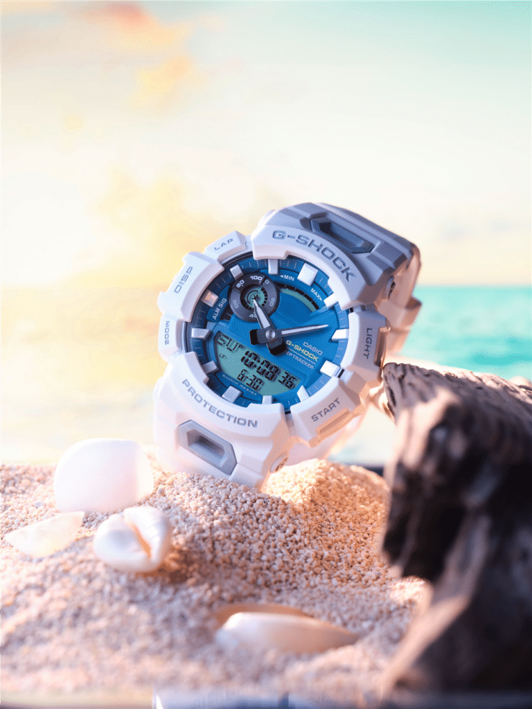 G-SHOCK watch in blue and white on sandy beach, evoking outdoor adventure and relaxation.