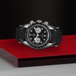 Tudor chronograph watch with black dial, stainless steel case, and leather strap on dynamic backdrop.