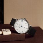 Elegant NOMOS wristwatch with leather strap and dominoes on a textured surface.