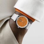 Stylish orange dial watch on a wrist with elegant sleeve and coordinated accessories.