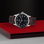Tudor watch with stainless steel case, black face, and brown leather strap on red background.