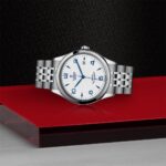 Elegant Tudor watch with stainless steel case, textured white dial, and blue accents on red platform.