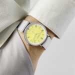 Stylish wrist watch with vibrant yellow dial and gray fabric strap against elegant clothing.