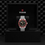 Luxurious Tudor watch in elegant box with black dial, red bezel, and stainless steel bracelet.