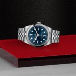 Elegant Tudor watch with stainless steel case and blue dial on a bold red platform.