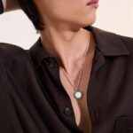 Sophisticated person in a dark brown shirt with a silver pendant necklace, exuding confidence.