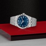 Luxurious stainless steel wristwatch with blue dial, diamond accents, and fluted bezel on red background.