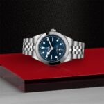 Tudor luxury watch on vibrant red background, showcasing blue dial and stainless steel bracelet.