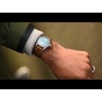 Stylish wristwatch with blue dial and leather strap, set against a tactical jacket background.