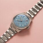 Stylish Oris wristwatch with blue dial and stainless steel bracelet against a pink backdrop.