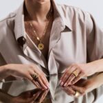 Elegant woman in a beige shirt showcases gold jewelry and confidence in a reflective portrait.