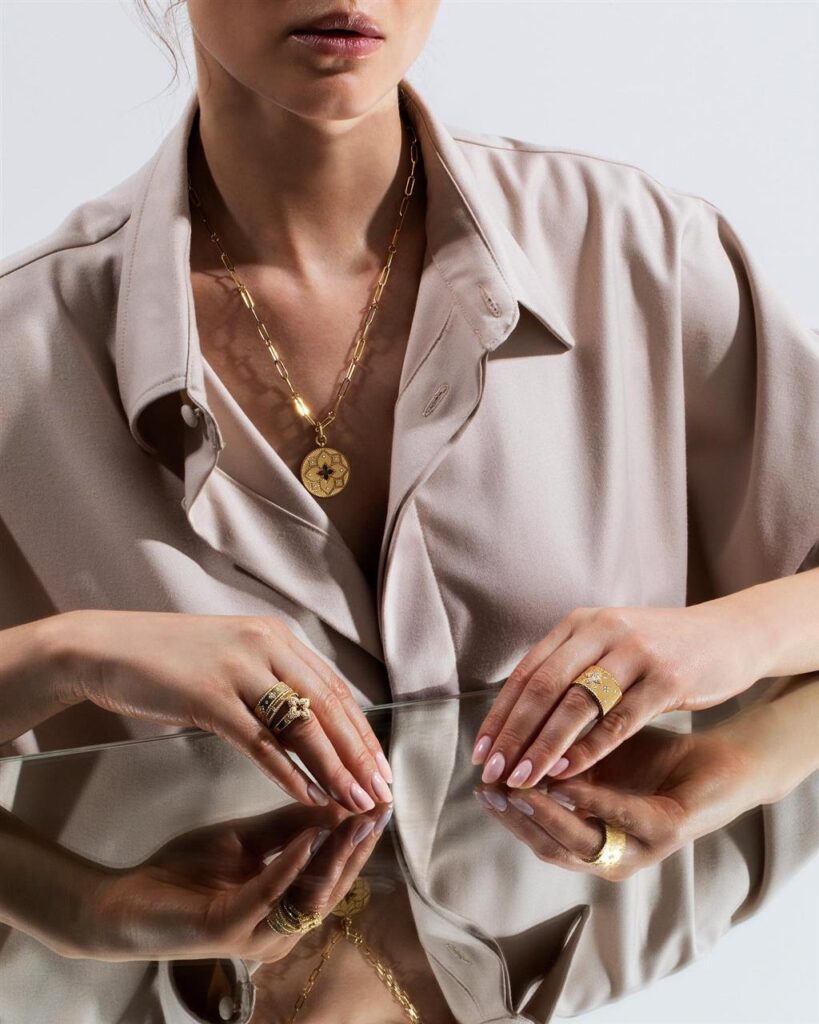Elegant woman in a beige shirt showcases gold jewelry and confidence in a reflective portrait.