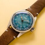 Stylish Oris automatic wristwatch with teal dial and brown leather strap on a warm backdrop.